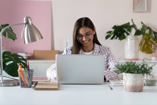 Happy young indian mixed-race business woman entrepreneur using computer looking at screen working in internet sit at office desk, smiling female professional employee typing email on laptop